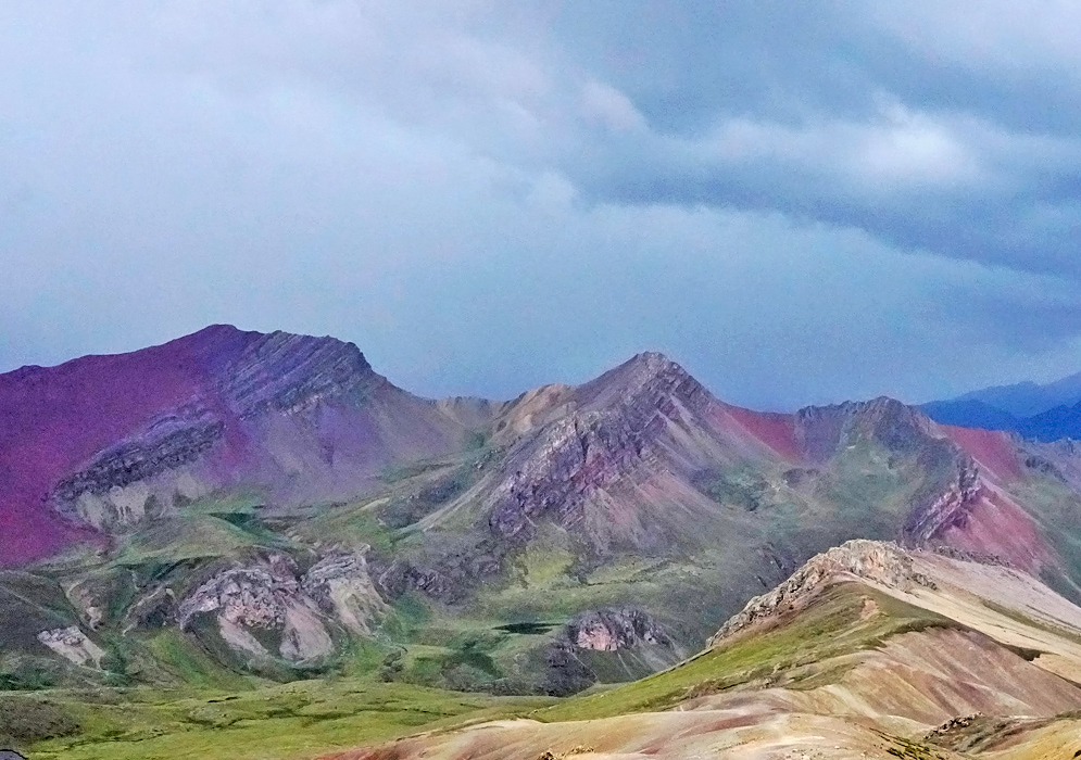 Jagged Andean peaks colored with stripes of red, purple, and olive green seen beneath an ominous stormy sky in the Rainbow Mountain range