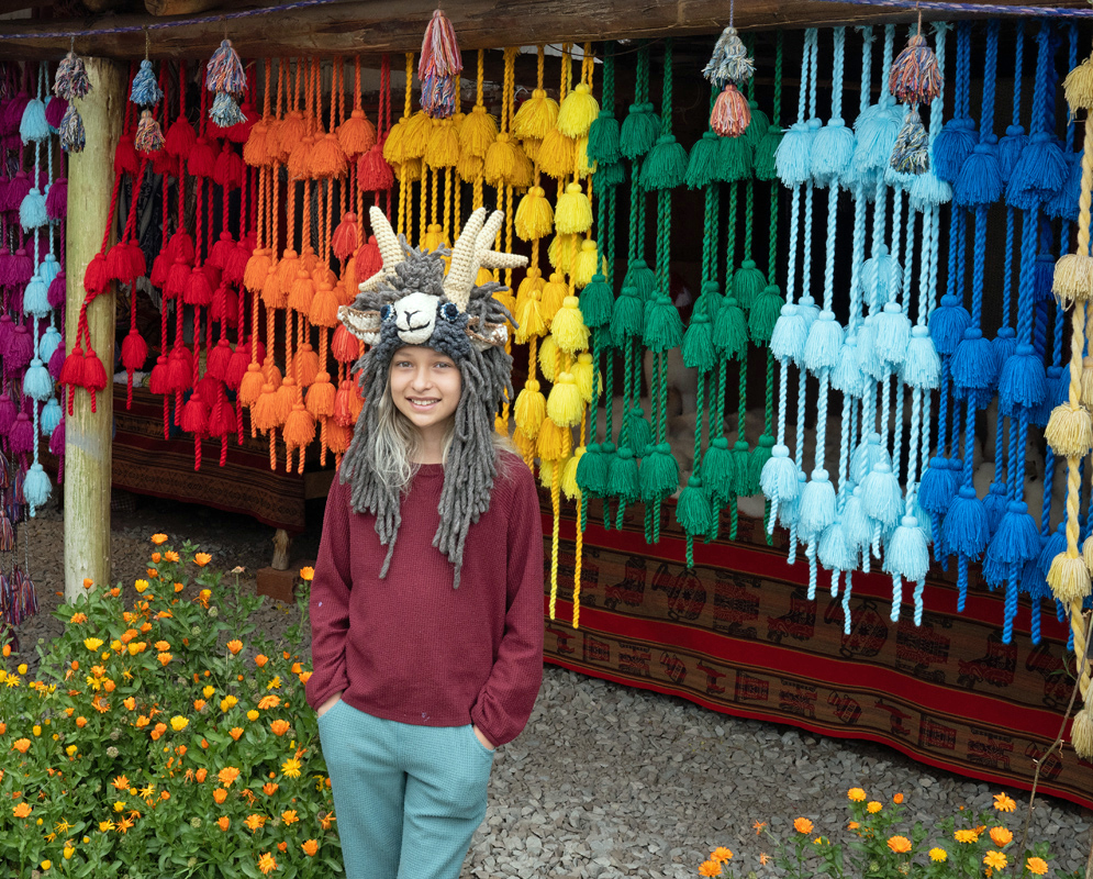 Girl with Handmade Hat in Front of Andean Tassels Smiling girl in animal hat standing in front of vibrant rainbow-colored Andean tassel garlands at a traditional Peruvian market