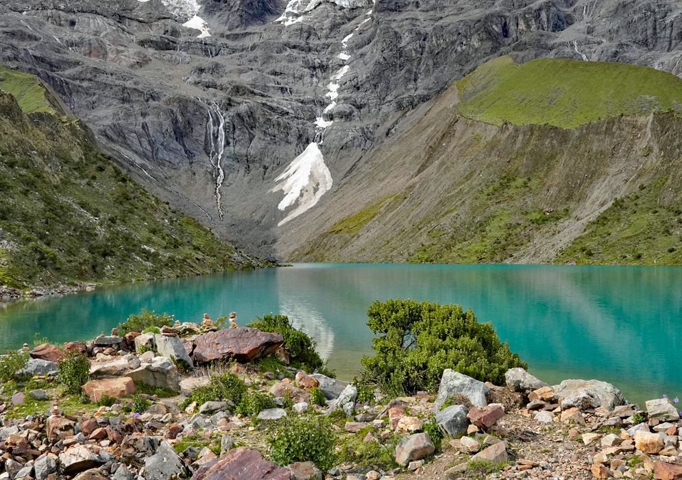 Humantay Lake Andes View The turquoise waters of Humantay Lake nestled beneath steep grassy hills and snowy cliffs of the Peruvian Andes with small rocky plants in the foreground