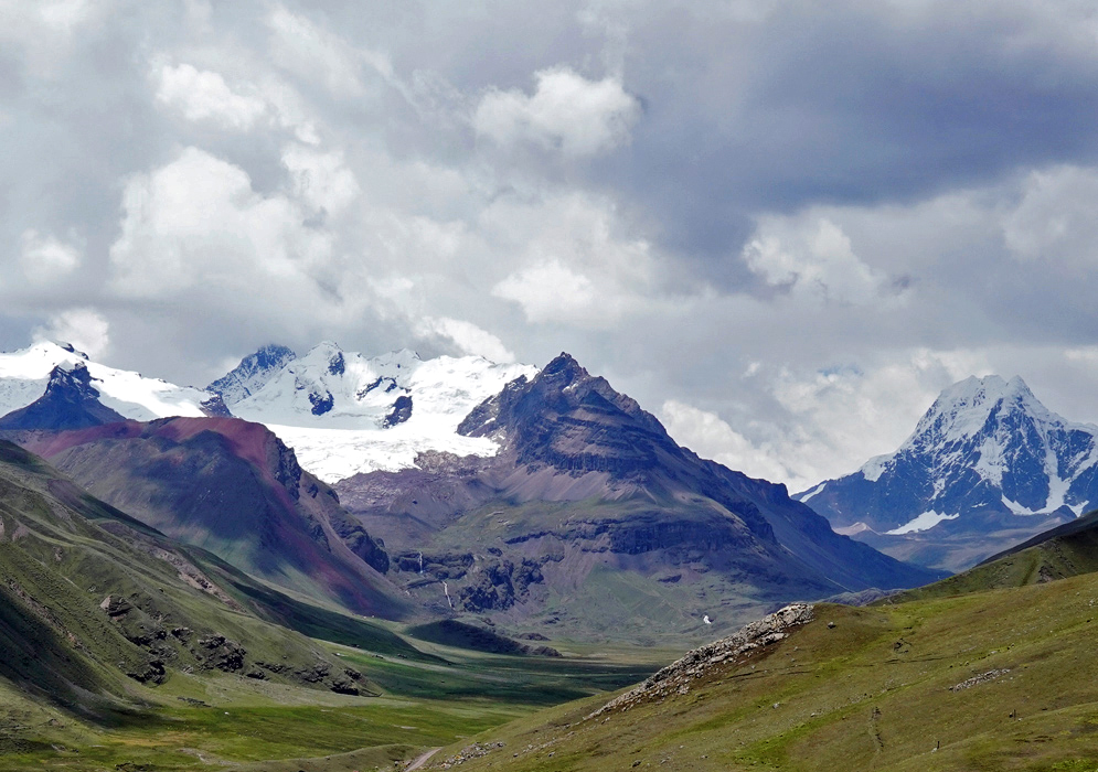 Snow-Capped Andes Peru Snow-capped peaks of the Peruvian Andes rise dramatically above a green high-altitude valley, with rugged cliffs and colorful mineral slopes under a cloudy sky