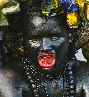 Carnival performer covered in glossy black body paint with red lips, bead necklaces, and a decorated headdress