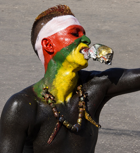 Carnival performer with red, yellow, green, and black body paint holding a small silver fish between his lips