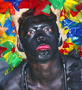 Carnival performer with a dark painted face, bright red lips, and a headdress made from multicolored ribbons