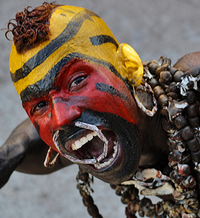 Carnival performer with red, yellow, and black face paint, prominent costume fangs, and layered shell jewelry