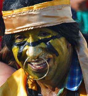 Smiling Carnival performer with gold and black face paint, a gold headband, and oversized hoop facial jewelry