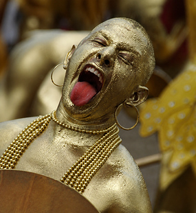 Carnival performer covered in metallic gold body paint singing while carrying a drum and wearing layered gold beads
