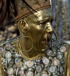 Carnival performer with metallic gold face paint, large hoop earrings, and an ornate costume covered in circular decorations