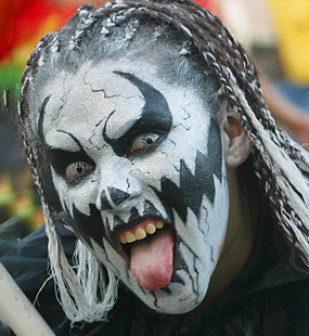 Carnival performer with dramatic black and white monster face paint, braided hair, and tongue extended