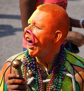 Carnival performer covered in orange body paint holding a wooden maraca and wearing colorful bead necklaces