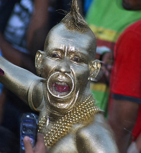 Carnival performer covered in metallic silver body paint with pointed hair, hoop earrings, and layered gold beads