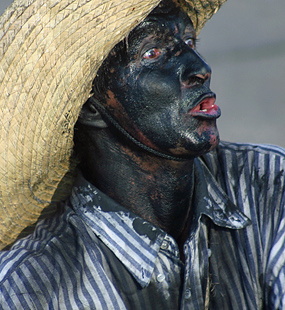 Carnival performer with a dark painted face wearing a wide straw hat and striped shirt while looking upward