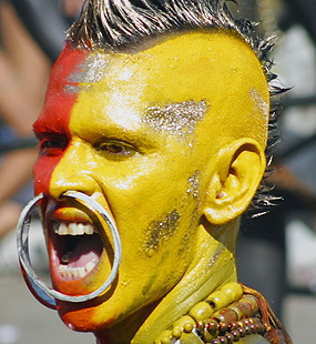 Carnival performer with vivid yellow and red face paint, a mohawk, large hoop jewelry, and an open-mouthed expression
