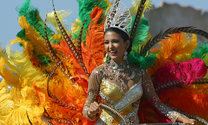 Barranquilla carnival queen in a sparkling gold dress and vibrant feathered costume, smiling and waving during the parade