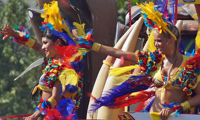 Two vibrant Barranquilla carnival dancers in colorful feathered costumes smiling and waving from a parade float