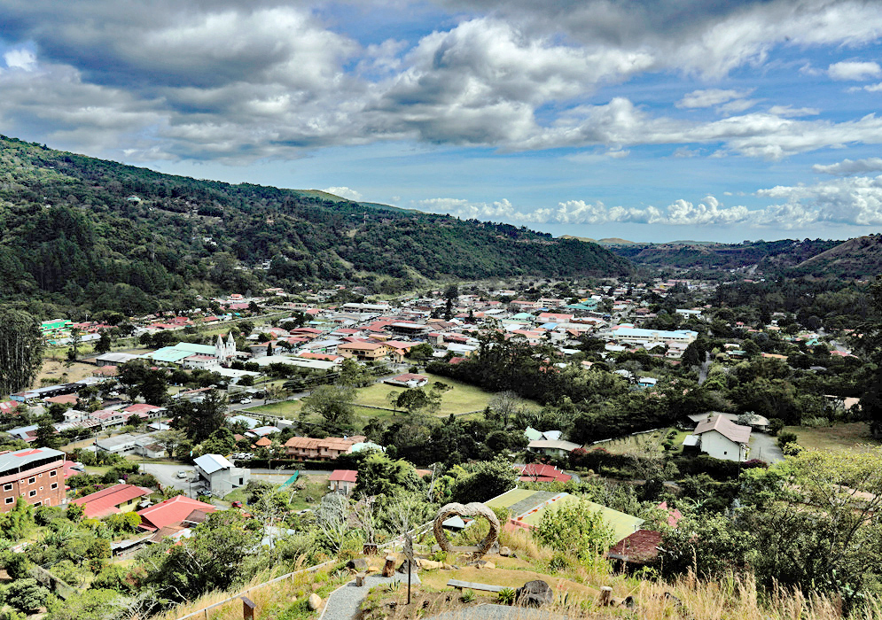 Boquete Panama Town Landscape Elevated view of Boquete, Panama, capturing the town nestled between green hills, with red-roofed homes, a white church, and a heart-shaped sculpture in the foreground under a partly cloudy sky