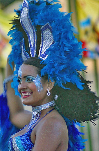 Barranquilla Carnival dancer in sparkling blue outfit and tall feathered headdress smiling during a colorful parade