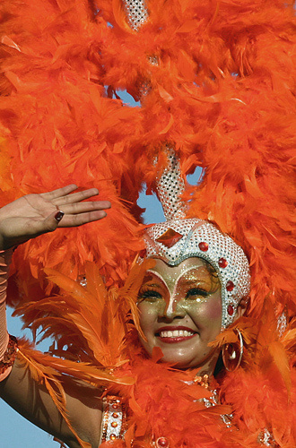 Carnival dancer in glittering silver headdress and vibrant orange feather costume waving and smiling at Barranquilla Carnival
