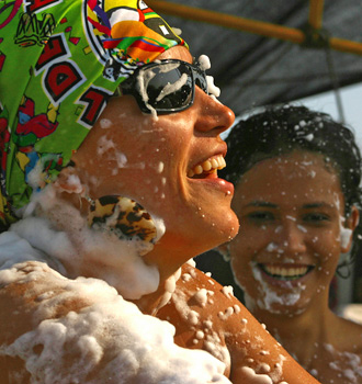 Karina laughs beneath a colorful Carnival hat as white foam covers her face, shoulders, and costume