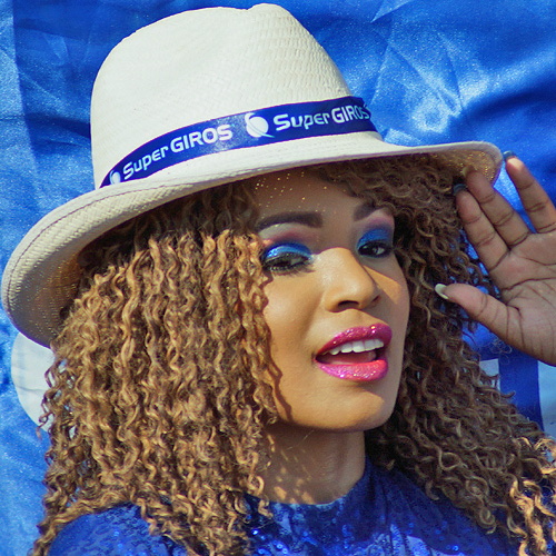Colombian carnival performer with curly hair, vibrant blue makeup, and a white Super GIROS hat smiling during Barranquilla Carnival