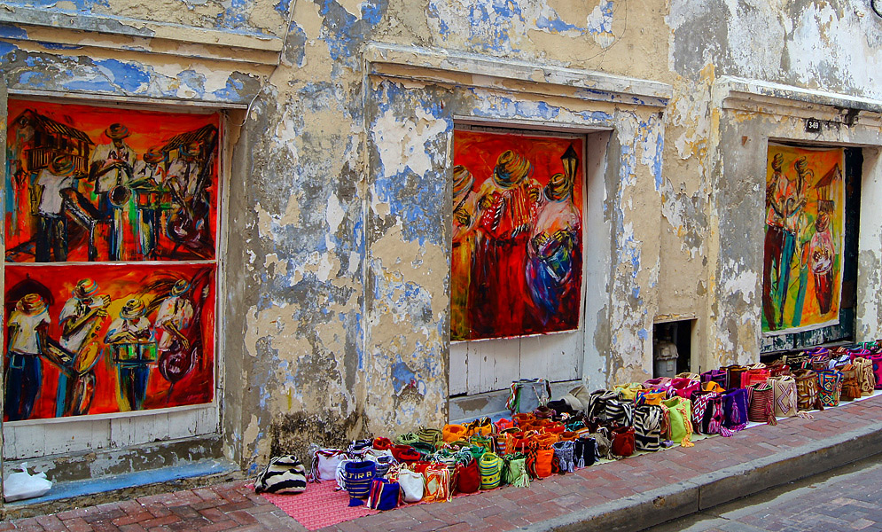 A sidewalk displaying colorful pursues and paintings in front of a deteriorating wall 