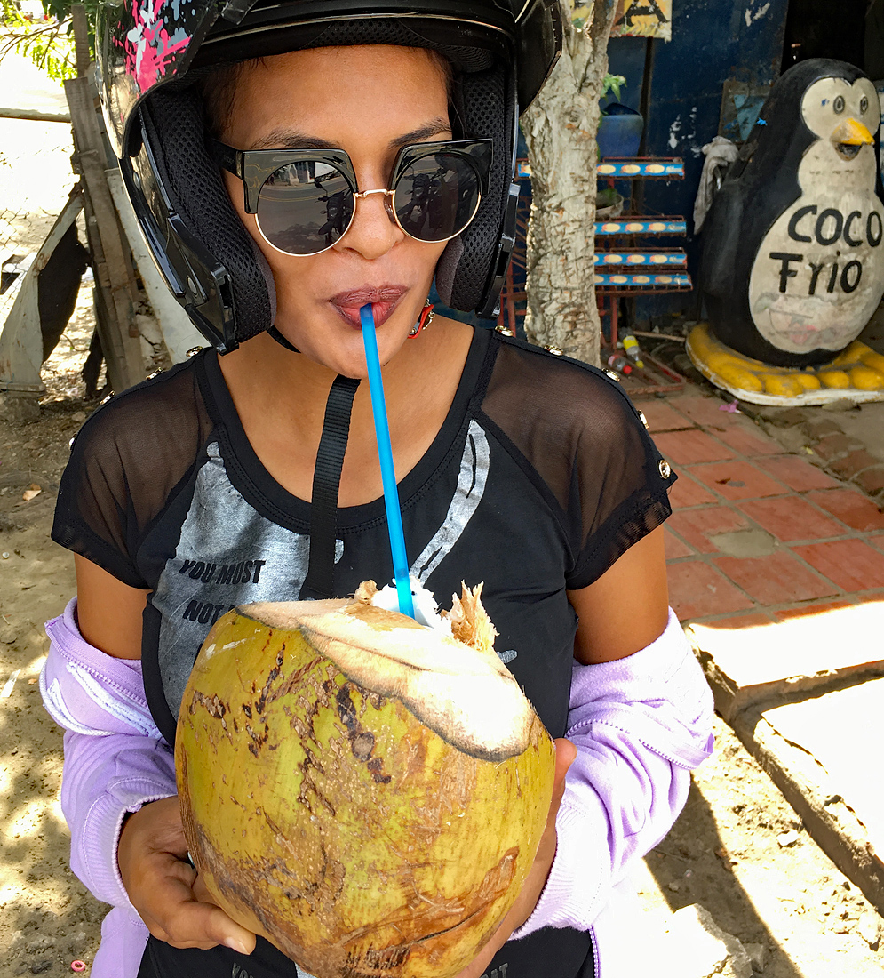 A Colombian woman wearing a motorcycle helmet and sunglasses drinking a Coco Frio with a straw from a large green coconut