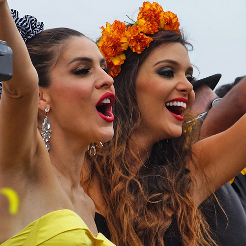 Two glamorous Colombian women with bright lipstick and floral headpieces cheering during the Barranquilla Carnival