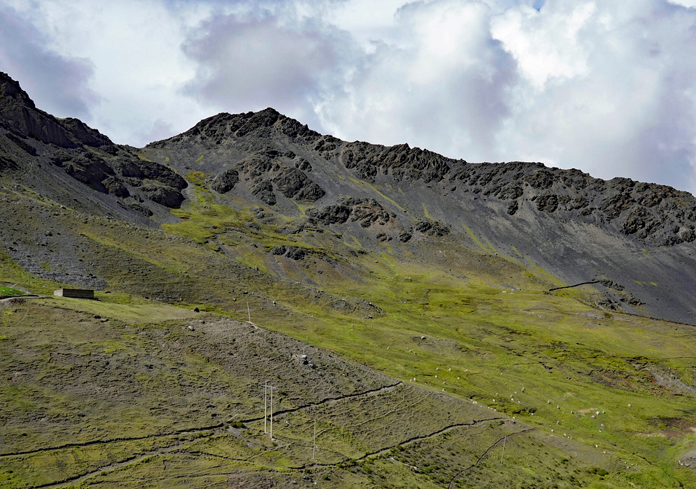 Cordillera Vilcanota Green Slopes Rugged black volcanic peaks rise above the rolling green tundra slopes of the Cordillera Vilcanota, with small scattered stone huts and distant grazing alpacas