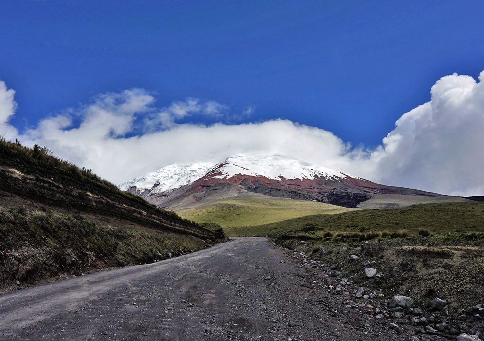 Road To Cotopaxi Volcano A dirt road leads directly toward the snow-capped Cotopaxi Volcano, with red mineral slopes and billowing clouds crowning the summit