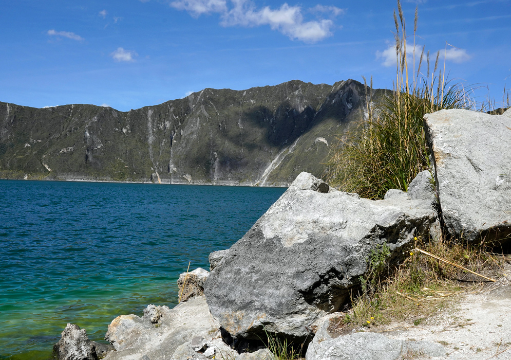 Quilotoa Lake Shoreline Close-up view of Quilotoa Lake’s shoreline with large gray volcanic rocks, tall grass, and the greenish-blue water beyond