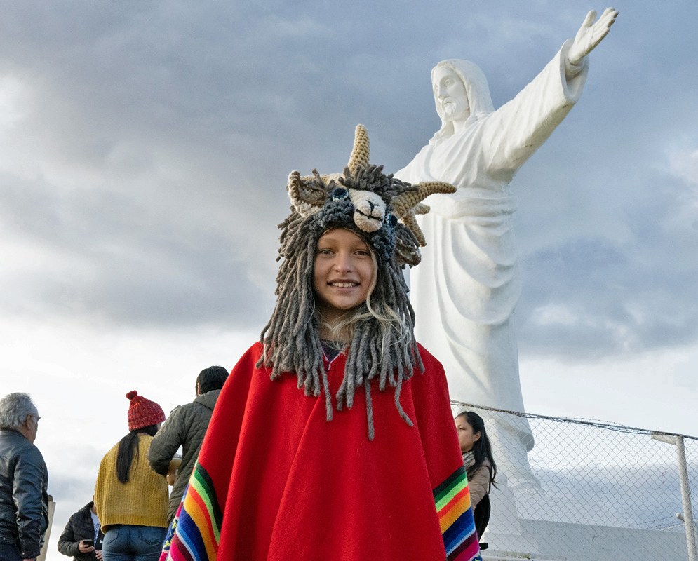 Cristo Blanco Statue in Cusco, Peru. Girl in red poncho and Andean hat standing below the Cristo Blanco statue overlooking Cusco, Peru