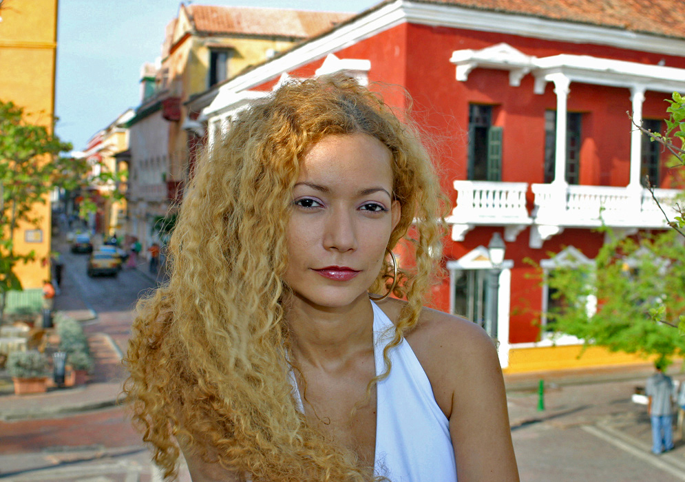 Colombian woman with long curly blonde hair wearing a white halter top with historic colorful colonial buildings in the background