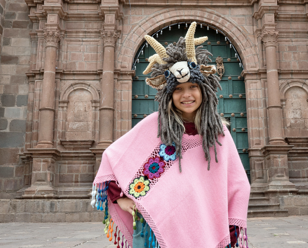 Andean Poncho and Historic Church Door in Cusco. Girl in pink Andean poncho and handmade animal hat smiling in front of a green church door in Cusco’s historic center