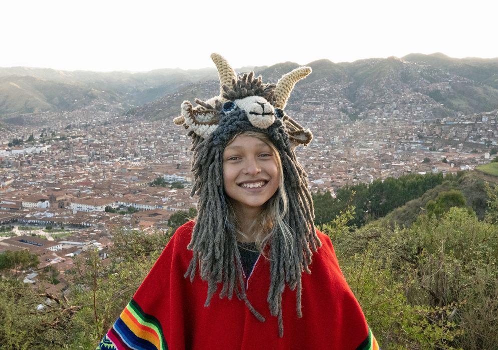 Smiling above Cusco with Andean Mountains in View Girl wearing colorful red poncho and ram hat smiling with panoramic view of Cusco, Peru and the surrounding Andes Mountains