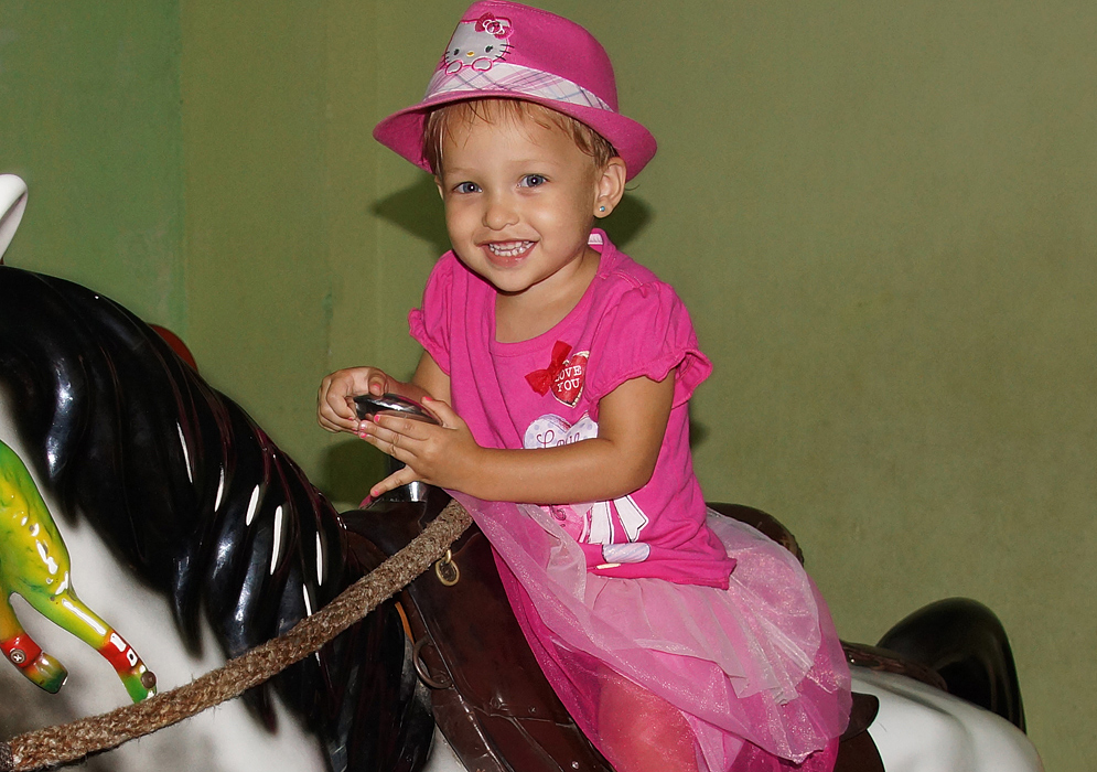 One-year-old girl with blonde hair and blue eyes in her pink outfit riding a toy horse