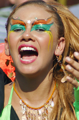 Singing Colombian woman with dramatic green and orange carnival face paint, golden earrings, and a vibrant expression shouting joyfully during Barranquilla Carnival