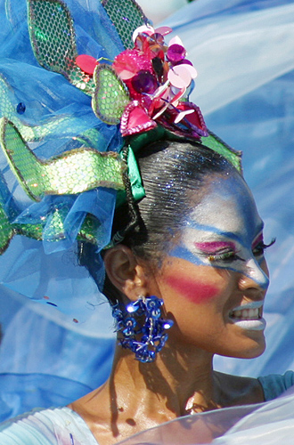 Colombian carnival dancer with blue and pink face paint, vibrant gemstone earrings, and a large feathered headpiece, performing with intense expression