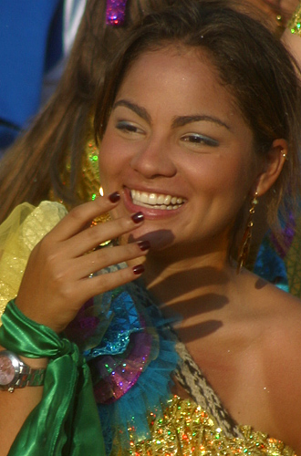 Smiling young Colombian woman in colorful carnival costume with glittering ruffles and green ribbon, caught mid-laughter while raising her hand