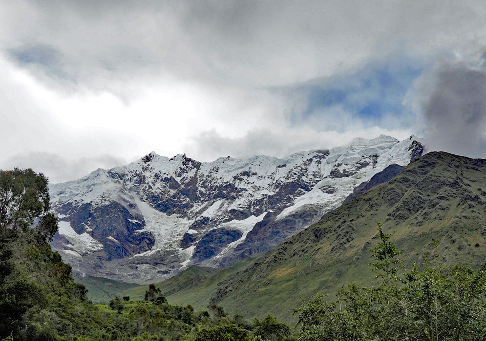 Humantay Andes Range The towering white-capped Humantay mountain range seen from a distance behind forested hills and soft green ridges beneath a dramatic cloudy sky