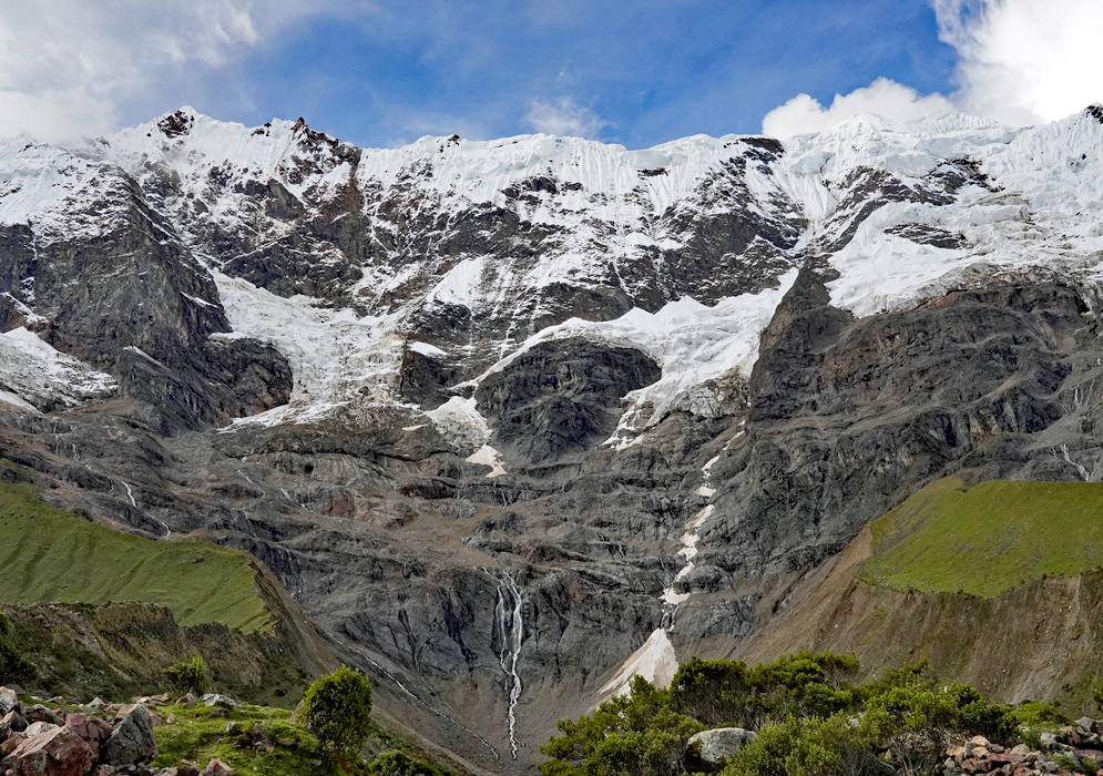 Humantay Mountain Glacier A dramatic snow-covered Andean peak with glacial meltwater cascading down rugged rock faces above lush green slopes near Humantay.