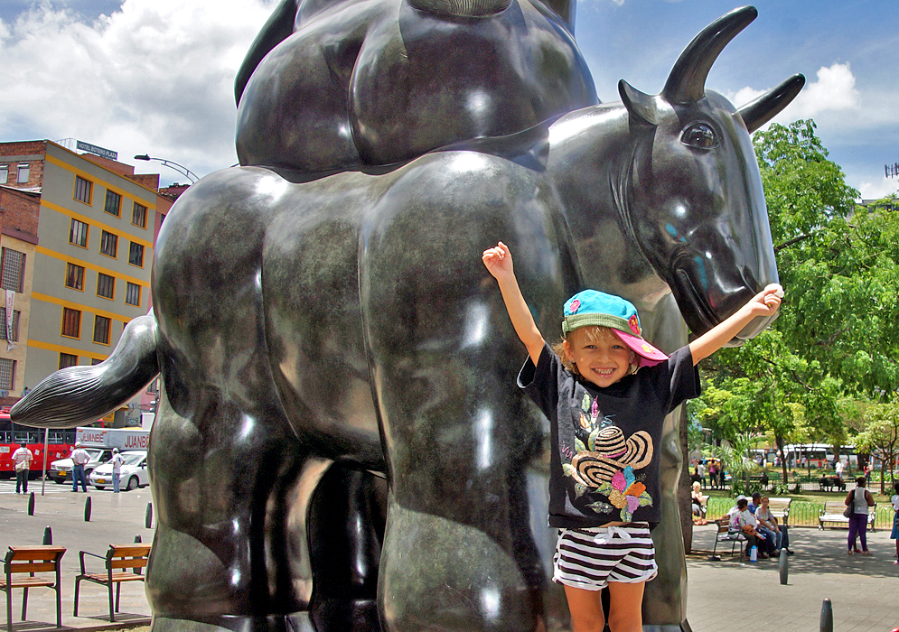 An attractive three year old holding her arms up in front of statue of a bull