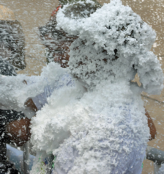  Person completely engulfed in thick white foam, with only faint facial features visible, during a playful foam battle at a carnival event