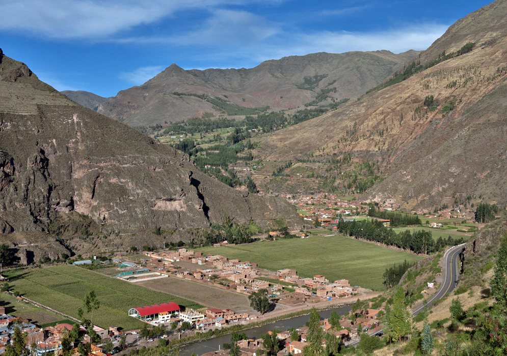 View of Ollantaytambo town and farmland extending across the valley floor, bordered by tall brown cliffs and stone terraces