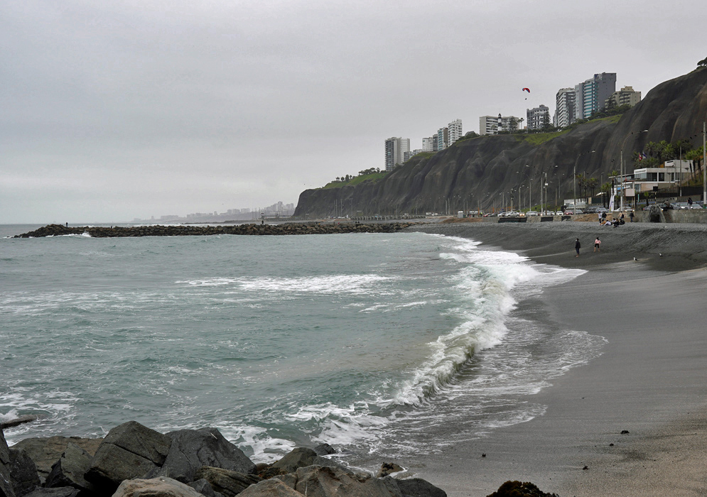 Pacific Coastline of Lima Rocky Pacific shoreline in Lima with gentle waves, dark cliffs topped by modern high-rises, and a cloudy coastal sky