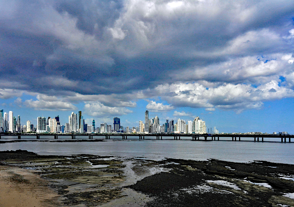 Panama City and Pacific Ocean View Broad view of Panama City’s skyline dominated by modern towers under dramatic storm clouds, with the Pacific Ocean and causeway in the foreground.