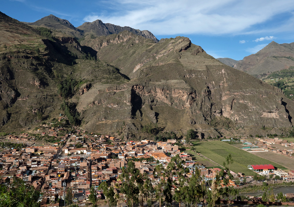 Distant view of Ollantaytambo framed by mountain walls and green cultivated lands, showing a cluster of red-roofed homes in the heart of the valley