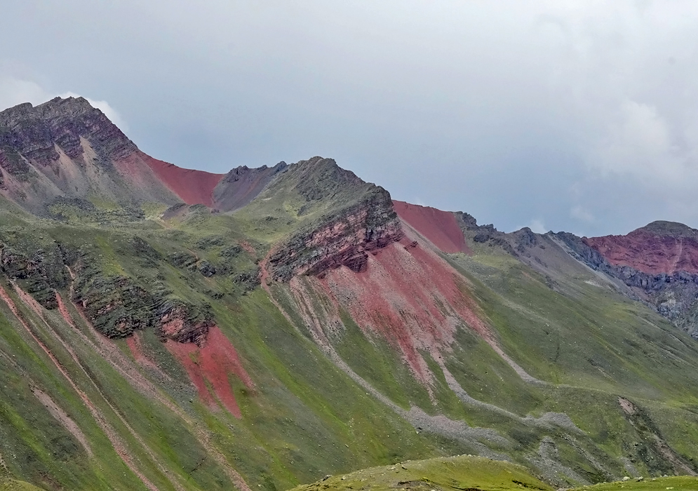 Steep Peruvian mountains with dramatic red mineral veins cutting through green slopes under an overcast sky in the Rainbow Mountains region
