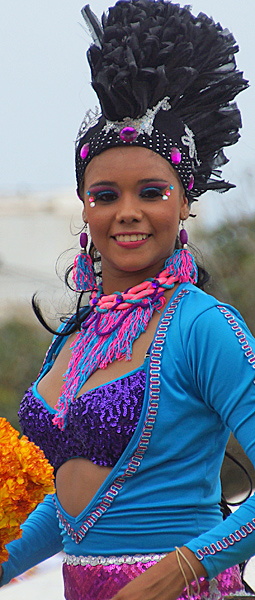 Barranquilla Carnival Dancer in Purple and Blue Colombian woman in a sparkling purple top, bright blue jacket, and tall feathered headdress, smiling during the Barranquilla Carnival parade with festive makeup and jewelry