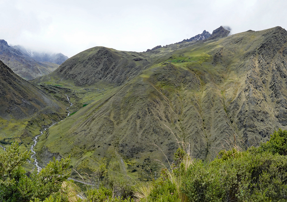 A wide mountain valley in the Cordillera Vilcabamba covered in high-altitude grassland and shrubs, with a small river cutting through the slope and fog hanging over the peaks