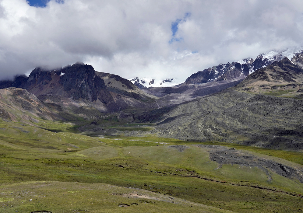 A panoramic view of the Cordillera Vilcanota’s glacier-capped peaks with layered dark volcanic rock and green highland plains below