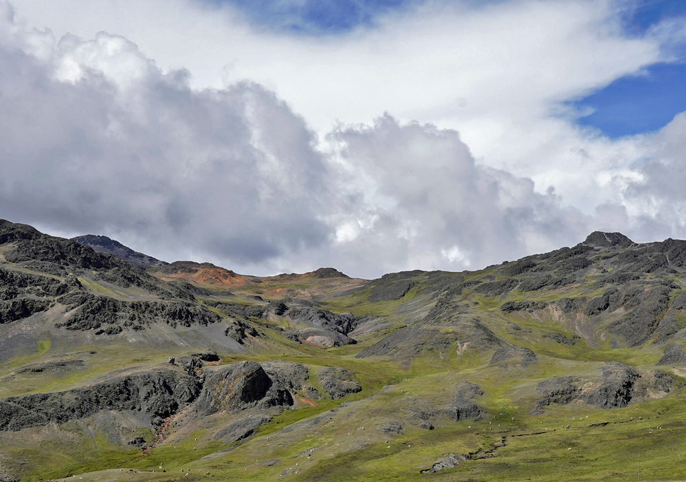 A mountainous landscape in the Vilcanota range with vibrant green slopes and steep rocky outcrops beneath a bright blue sky with scattered clouds